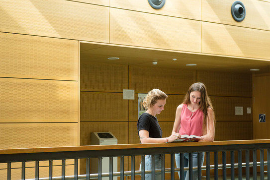 Studentinnen mit Buch am Geländer Zwei Studentinnen stehen mit einem Buch am Geländer.