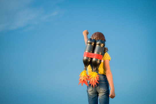 Man sieht eine junge Person mit gelbem T-Shirt, Jeans und Lederkappe von hinten vor einem blauen Himmel. Sie hat eine Attrappe eines Raketenrucksacks auf dem Rücken und streckt die linke Faust in die Höhe.